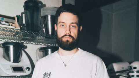 Portrait of a bearded person wearing a white T-shirt and necklace, standing in a kitchen or food-preparation area. Metal wire shelves behind the person hold several countertop appliances, mixing bowls, and containers. The image is lit with direct flash, creating strong shadows on the wall.