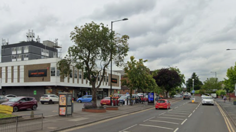 A road heading off into the distance with cars on it. On the left is a large shopping centre with cars parked outside
