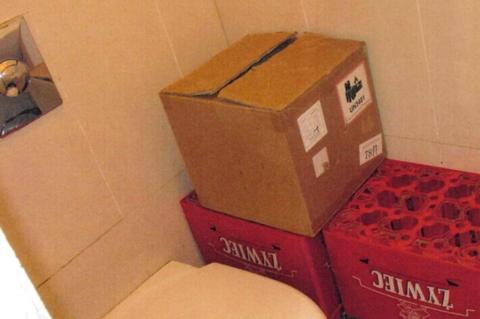 A brown box of fake cigarettes located next to a toilet seat. The box is perched on top of two beer crates.