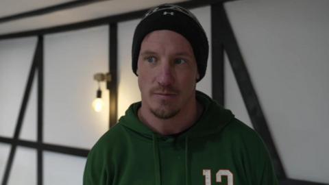 Billy Vernon stood in a room of his house. he is wearing a dark green hoodie and a black beanie hat. 