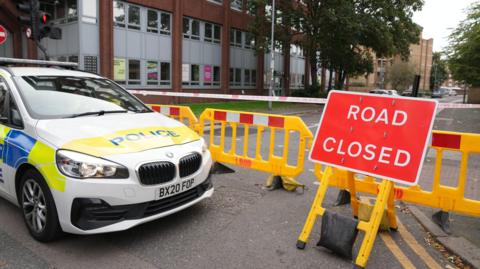 A police car parked in front of police cordon tape and a yellow barrier. A sign reading road closed is also in place.