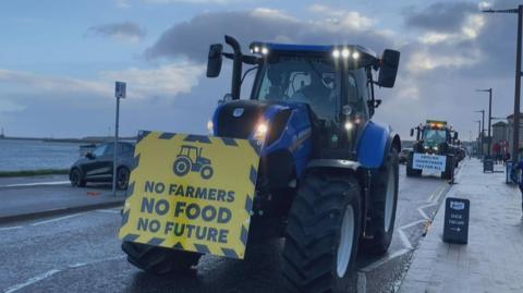 A blue tractor with a yellow sign on the front which says "No farmers, no food, no future". A green tractor is behind it, with a sign which says "abolish inheritance tax for all". The North Sea is in the background of the image. 