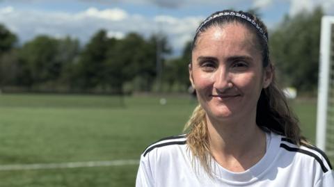A head and shoulders image of a young woman wearing a white and black football shirt. She is standing on a football pitch with a goal behind her. She is smiling and looking into the camera.