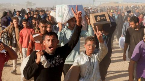 A Palestinian man and boy in the foreground look upset and make the 'peace' sign while carrying aid, including a box branded "GHF" for the Gaza Humanitarian Foundation. 