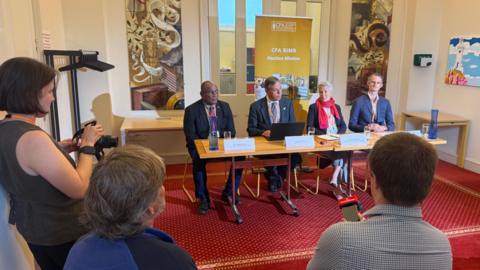 A woman and three men sitting behind a table, in front of a yellow banner which says 'CPA BIMR Election Mission' in white writing. In the foreground are three members of the press, holding cameras and microphones. The carpet is maroon with yellow / gold detailing.