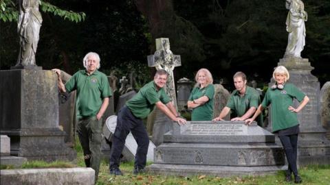 A group of people in green branded polo shirts stand amidst gravestones and memorial statues in the cemetery. Three people are leaning on a large stone memorial.