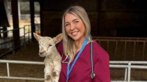 Karis Ramsay holding a lamb in front on an animal pen. Karis is wearing burgundy scrubs, a blue lanyard and ID as well and a stethoscope 