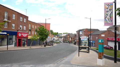 A row of shops with a road in the centre. A bus stop is on one of the footpaths and there are artistic pink signs reading Wakefield attached to the lamp posts.