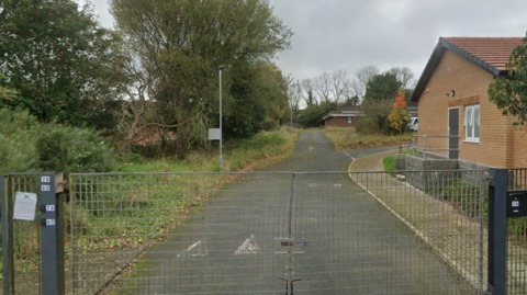 A locked metal gate is closed across a quiet tarmac path leading past a brick building on the right and overgrown greenery and trees on the left, under a cloudy sky.