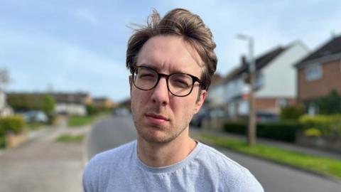 Oli Franklin-Wallis looks at the camera with his eyes squinting and not smiling. He is standing in a residential road wearing a grey T-shirt and glasses. He has windswept hair.
