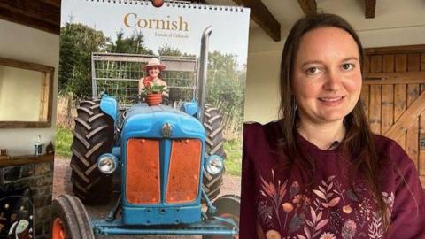 Jessica Riggs, on the right, is wearing a maroon jumper and has shoulder length brown hair. She is holding a calendar which she has made, which has an image of her wearing a pink hate on a blue tractor. 