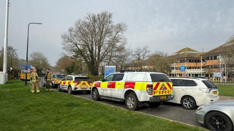 Fire crews outside Weston hospital on Thursday. A line of Fire Service vehicles line the road, with fire officers huddled on a grassy verge speaking to one another.