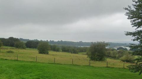 A set of green fields with a fence running through the middle. There are trees in the distance and on the horizon the skyline of a city can be seen.