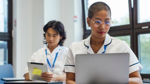 A student nurse sits looking at a laptop in a classroom. She has short cropped her and wears gold-rimmed glasses.