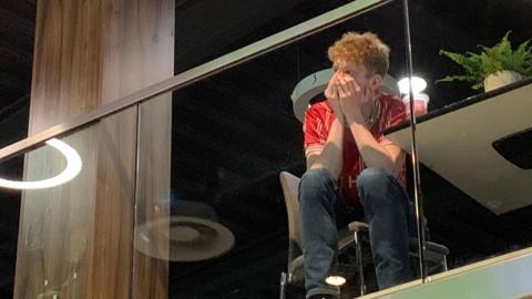 A man sat on a chair behind a glass screen at the sport bar at Ashton Gate. He is wearing jeans and a red Bristol City shirt. His hands are in his face as he looks on.