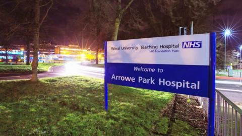 A night-time view of a blue sign saying 'Welcome to Arrowe Park Hospital" in the foreground with the lights of the hospital building shining in the background