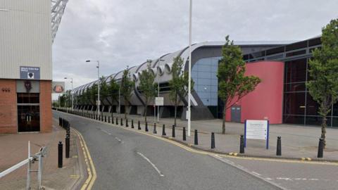 Sunderland Aquatic Centre stands to the right of the Stadium of Light. The Aquatic Centre has a large curved wall which along the length of the road. It is grey with a red wall near its entrance. The side of the Stadium of Light can be seen to the left. A sign with the club's badge is hanging from the wall while a door is marked with a black cat emblem.