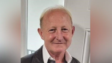 Scott Lisney smiles at the camera. He wears a leather jacket with a shirt underneath that has a large white collar. 