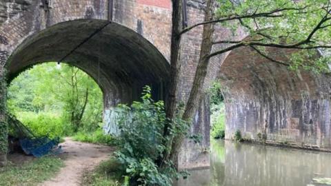 A photo looking under two arches of a bridge over the River Frome and a pathway alongside it which is surrounded by grass and overgrown vegetation. 