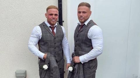 Corey and Liam Beavis wearing matching grey suits and white shirts, with burgundy tie and grey waistcoat. They have short light brown hair.