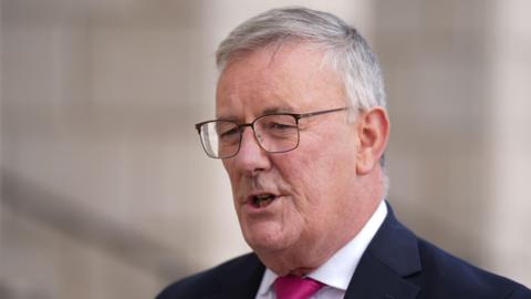 Mike Nesbitt has short grey hair and metal square glasses. He is speaking. He is wearing a white shirt, pink tie and a black blazer.