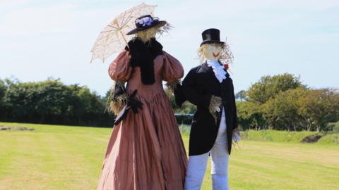 A picture of two scarecrows holding hands at a previous event. One is wearing a black and white suit and one is wearing a red dress. 