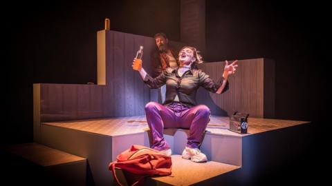 A theatre show. A woman in purple trousers and a brown top sits on stage holding a bottle of orange liquid. Behind her a man is leaning against a wall