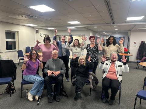 A group of nine people, four seated in front of nine standing, all waving their hands in the air and smiling.