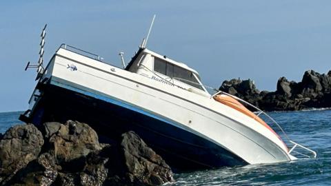 A white boat is balancing on the top of rocks. Part of the nose is leaning into the water.