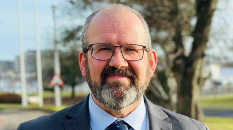 Mark Findlater, wearing glasses and a shirt, jacket and tie, with trees in the background.