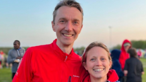 Mike Green stands with his arms around his wife Collette. They are both smiling at the camera. They are standing outside in a sports pitch area with people behind them. Mike has short light hair and wears a red running jumper. Collette has light hair and wears a red hoodie with a coat over the top.