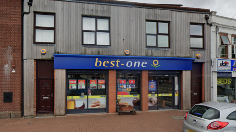 A shop front. The shop is the ground floor of a brick building with wood over the bricks. It has blue hoarding above with best written in yellow and one written in while. 