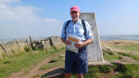 Andrew Simpkins smiles at the camera, standing on the stop of a hill with a lovely vistal in the background of rolling hills and a blue sky. He's wearing a red cap and a light blue shirt, and holding a plastic wallet with maps and documents inside.