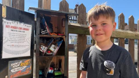 A young boy smiles to camera wearing a grey t-shirt. To his left is a birdbox style toy swap box full of toy cars.