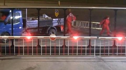 Still from CCTV shows two women in red overalls carrying a silver ladder behind a large security fence. Their associate is sitting in the cab of a blue flatbed truck next to them with a pile of rubble on its back.