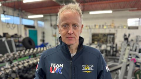 Rob Shenton is photographed in profile at a gym. He has wispy, short grey hair and is wearing a branded Team UK Invictus Games blue tracksuit top. Rows of free weights and exercise bikes can be seen in the background.