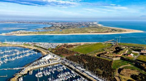 An aerial photo shows the shoreline at Eastney with yachts moored in the harbour. In the distance you can see the Hayling Island peninsular with golden sands. The sea is as deep blue. 