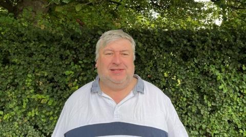 Councillor John Hubbard stands in front of a hedge while wearing a light grey shirt. He has short grey hair and grey stubble.
