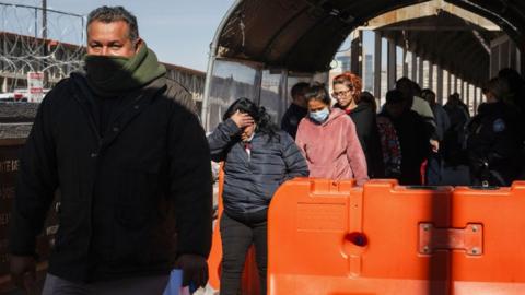 A group of people walk towards the camera, with their faces partially covered. They are walking past a large orange barrier