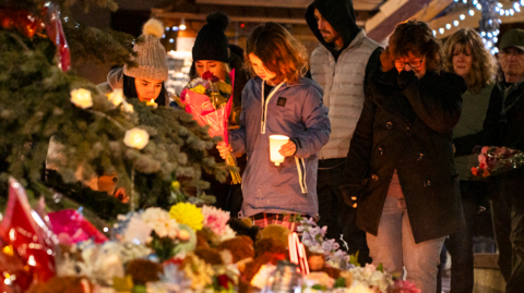 Community members mourn during a candlelight vigil for the victims of Tumbler Ridge Secondary School where a mass shooting took place a day earlier in the small town of Tumbler Ridge, British Columbia, on February 11, 2026. A pile of flowers and candles can be seen at the forefront of the image, while several people look down in the background towards the vigil with sombre expressions including a young girl in the centre of the image