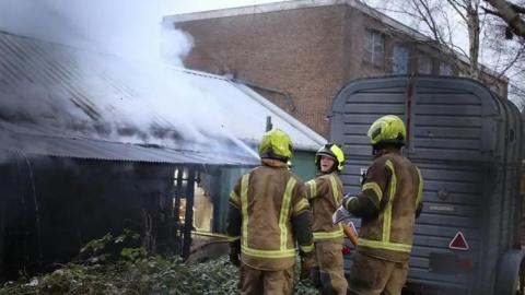 Three firefighters in full protective gear are positioned beside a metal‑roofed building. Thick white smoke is pouring out from the roof.
