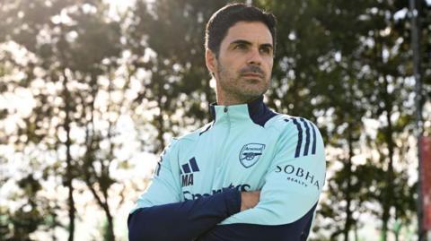Mikel Arteta stood with his arms folded looked away from the camera against a backdrop of trees wearing a light blue arsenal training top.