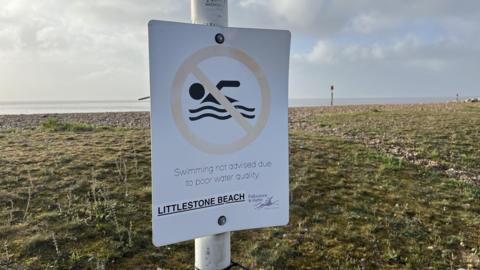 A sign on a lampost with the outline of a swimmer in waves, circled in red with a line through the middle. The sign reads "Swimming not advised due to poor water quality" and "Littlestone beach". A shingle beach is seen in the background.