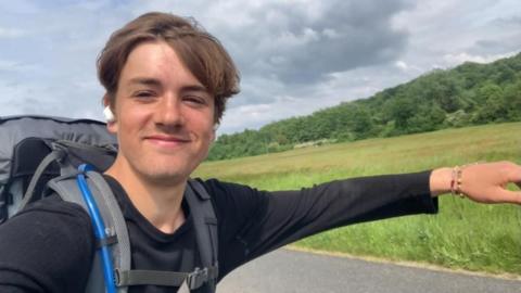A young man skateboards along an open road lined by long grass and trees. The skateboard is not visible but he has his arms outstretched to balance himself
