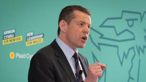 Rhun ap Iorwerth, a man in a dark suit, blue shirt and burgundy tie, speaks on stage.