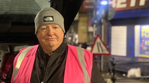 Ronny English wearing a grey The North Face hat and pink hi-vis jacket over a black jacket in front of a vehicle with resources. 