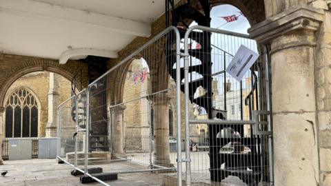 Metal fencing positioned in the arched open area under Peterborough's Guildhall. It conceals to winding staircases. A church is behind the Guildhall.