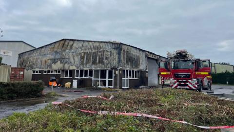 Image shows partially collapsed warehouse at a Plympton business park. The building is partially collapsed. A fire engine is parked next to the building. 