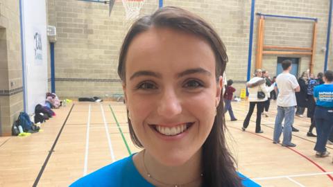 Leah is wearing a blue t-shirt, she has long brown hair and a small chain around her neck. She is standing in a busy sports hall with parents and children behind her.