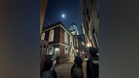 Three men stand outside the synagogue at night and point towards the moon, which appears full. 